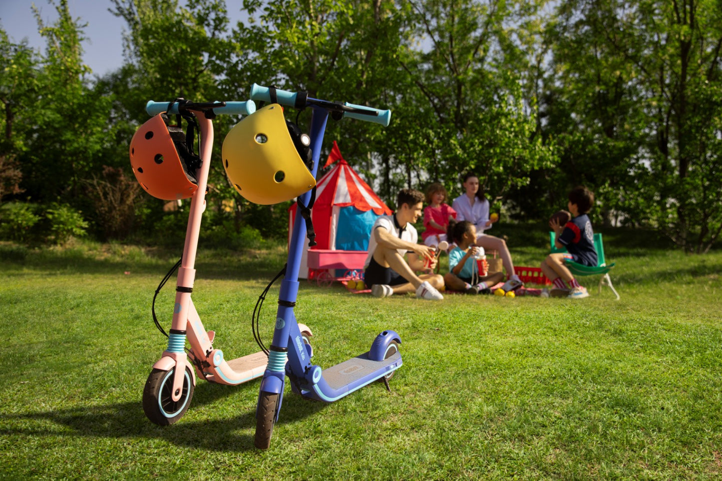 Family picnic on a sunny summer day. Two Segway Ninebot Zing e8 electric kickscooters (one pink, one blue) in the foreground. Helmets hanging off of the handlebars. 